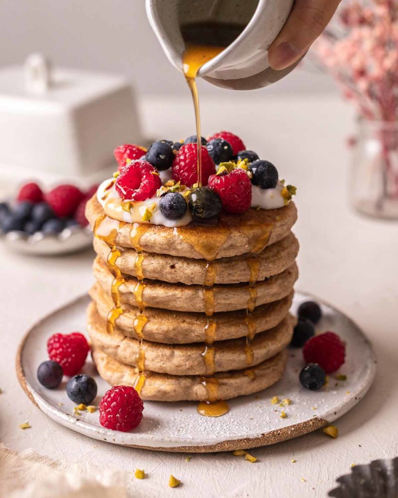A stack of 6 fluffy whole grain pancakes on a rustic ceramic paint. Fresh raspberries and blueberries are scattered around the pancakes on the plate, and the stack of pancakes is topped with a creamy topping, more fresh berries, and chopped pistachios. At the top of the picture, a hand is holding a small, white ceramic pitcher from which the person is drizzling maple syrup over the stack of pancakes.